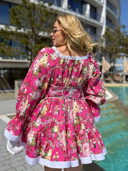 A woman wearing a bright pink floral long-sleeve dress with white ruffle trim, photographed outdoors near a swimming pool and modern building.