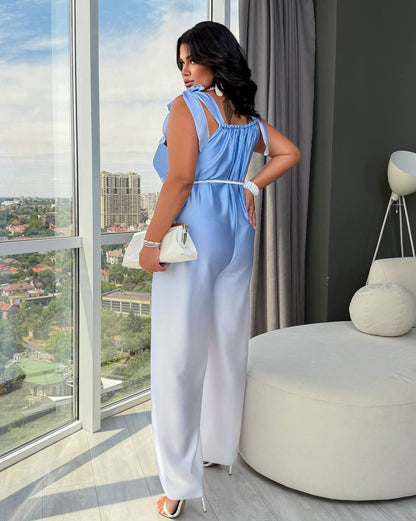 A woman stands by a large window in a modern room, wearing a sleeveless light blue and white jumpsuit with a tie waist, holding a small white handbag.
