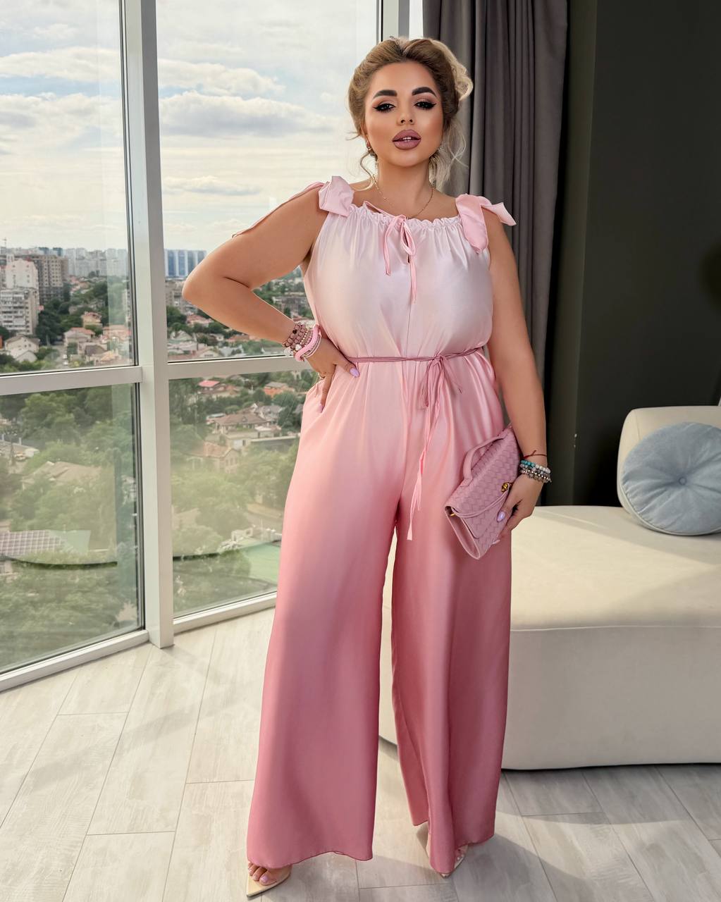 A woman models a light pink satin-like jumpsuit with ruffled shoulder straps, a tie neckline, a matching waist tie, and wide legs. She is standing in a modern room with a large window showing a city view, holding a matching pink clutch.