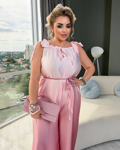 A woman models a light pink satin-like jumpsuit with ruffled shoulder straps, a tie neckline, and a matching waist tie. She is holding a matching pink quilted clutch in a modern living room setting with a large window overlooking a cityscape.