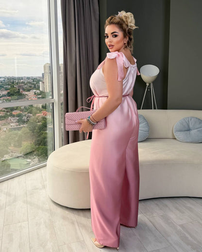 A woman models a light pink satin jumpsuit with ruffled shoulder straps and a tie waist, holding a matching pink quilted clutch in a modern living room setting.