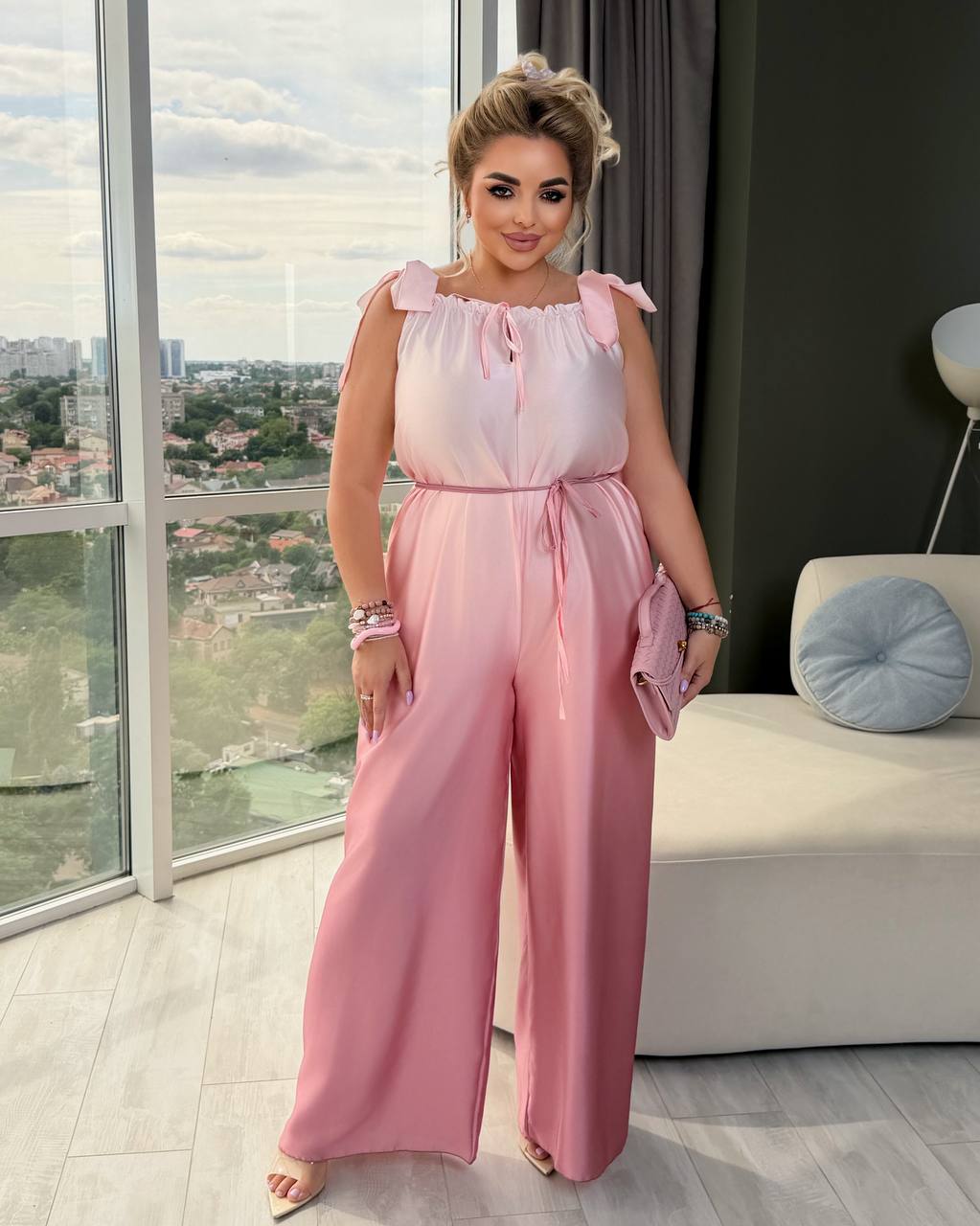 A woman modeling a light pink wide-leg jumpsuit with ruffled shoulder straps and a tie waist, standing in a modern room with a large window overlooking a cityscape.