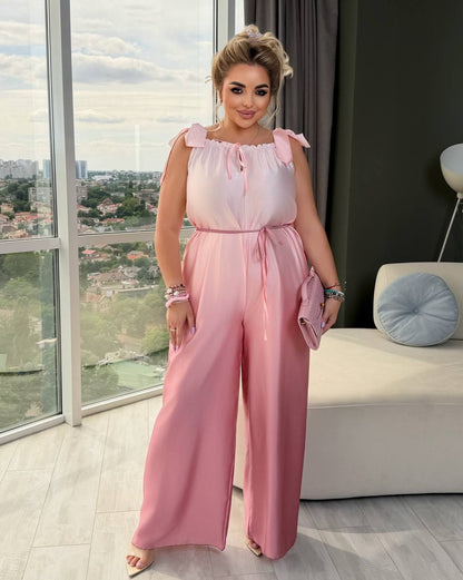 A woman modeling a light pink wide-leg jumpsuit with ruffled shoulder straps and a tie waist, standing in a modern room with a large window overlooking a cityscape.