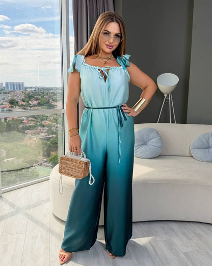 A woman models a sleeveless gradient jumpsuit with ruffled shoulders and wide legs, transitioning from light aqua to deep teal. She is standing in a modern living room with a large window showing a city view, holding a small woven handbag.