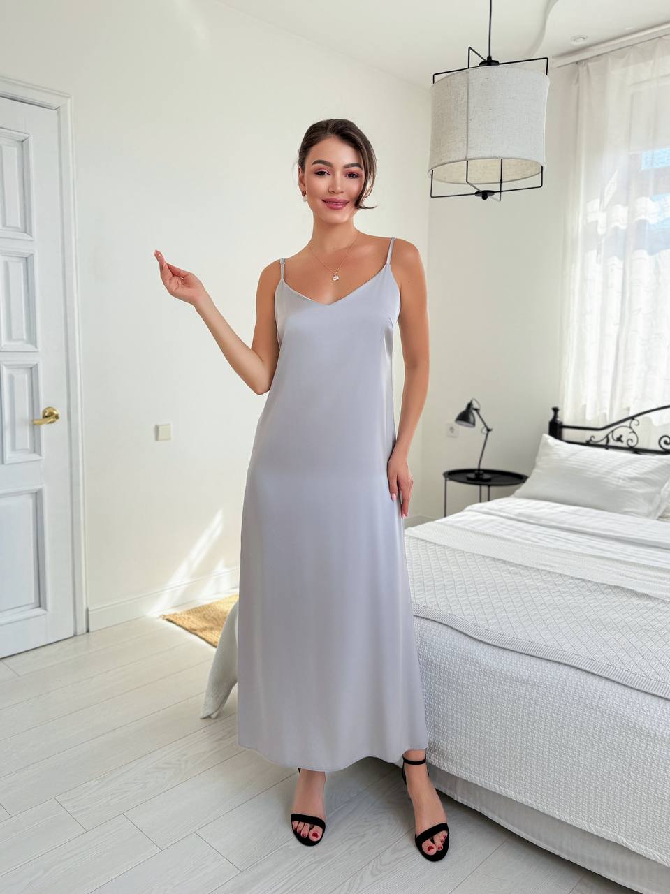 A woman modeling a light gray slip dress in a bright, modern bedroom setting with white walls, a bed, and a hanging pendant lamp.