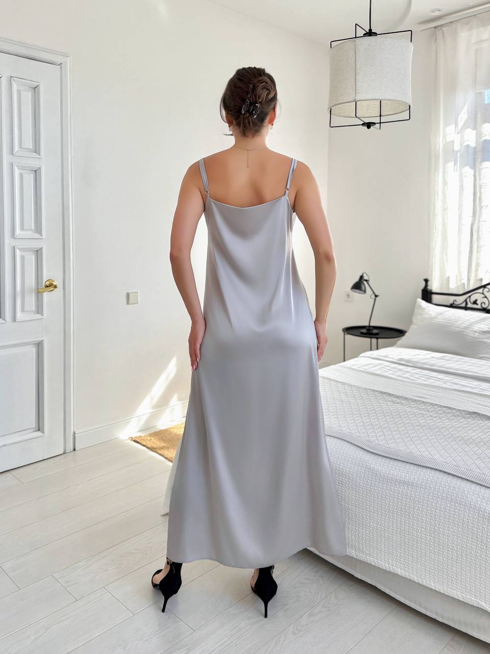 A woman viewed from behind wearing a light gray satin slip dress in a bright, modern bedroom with white walls, a bed, and a hanging pendant light.