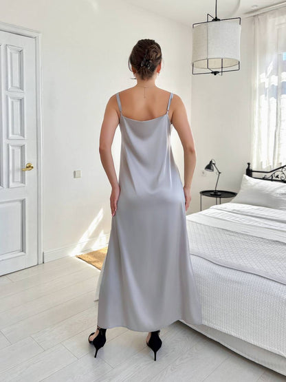 A woman viewed from behind wearing a light gray satin slip dress in a bright, modern bedroom with white walls, a bed, and a hanging pendant light.