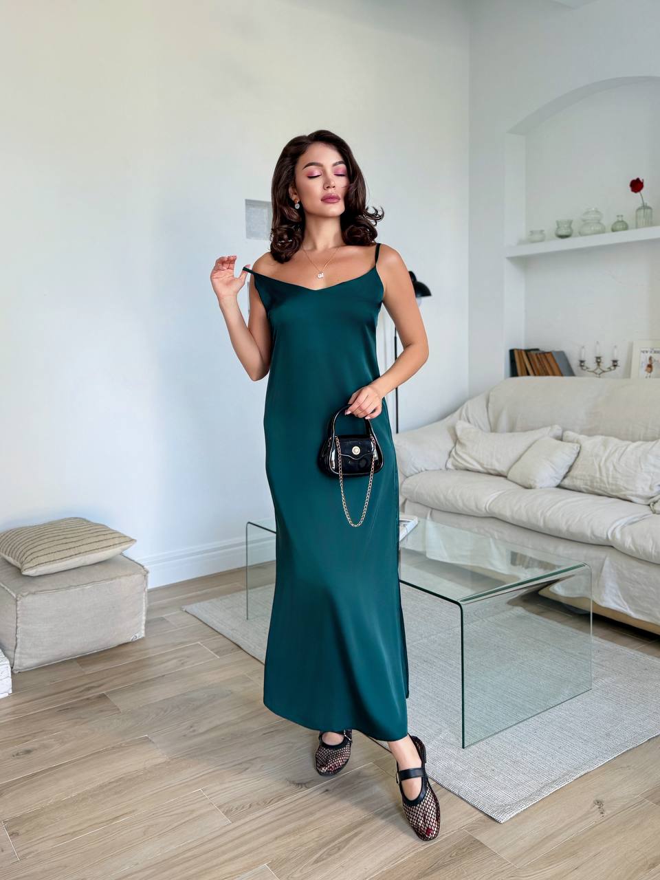 A woman wearing a dark teal green spaghetti-strap maxi dress, holding a small black handbag, standing in a modern living room with light wood floors and a white sofa.