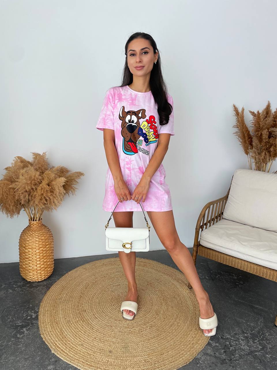 A woman stands indoors modeling a pink tie-dye t-shirt dress with a colorful cartoon graphic on the front. She is holding a white handbag and wearing white slide sandals, posed on a round woven rug next to a sofa and pampas grass decor.