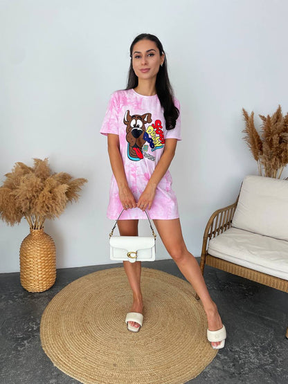A woman stands indoors modeling a pink tie-dye t-shirt dress with a colorful cartoon graphic on the front. She is holding a white handbag and wearing white slide sandals, posed on a round woven rug next to a sofa and pampas grass decor.