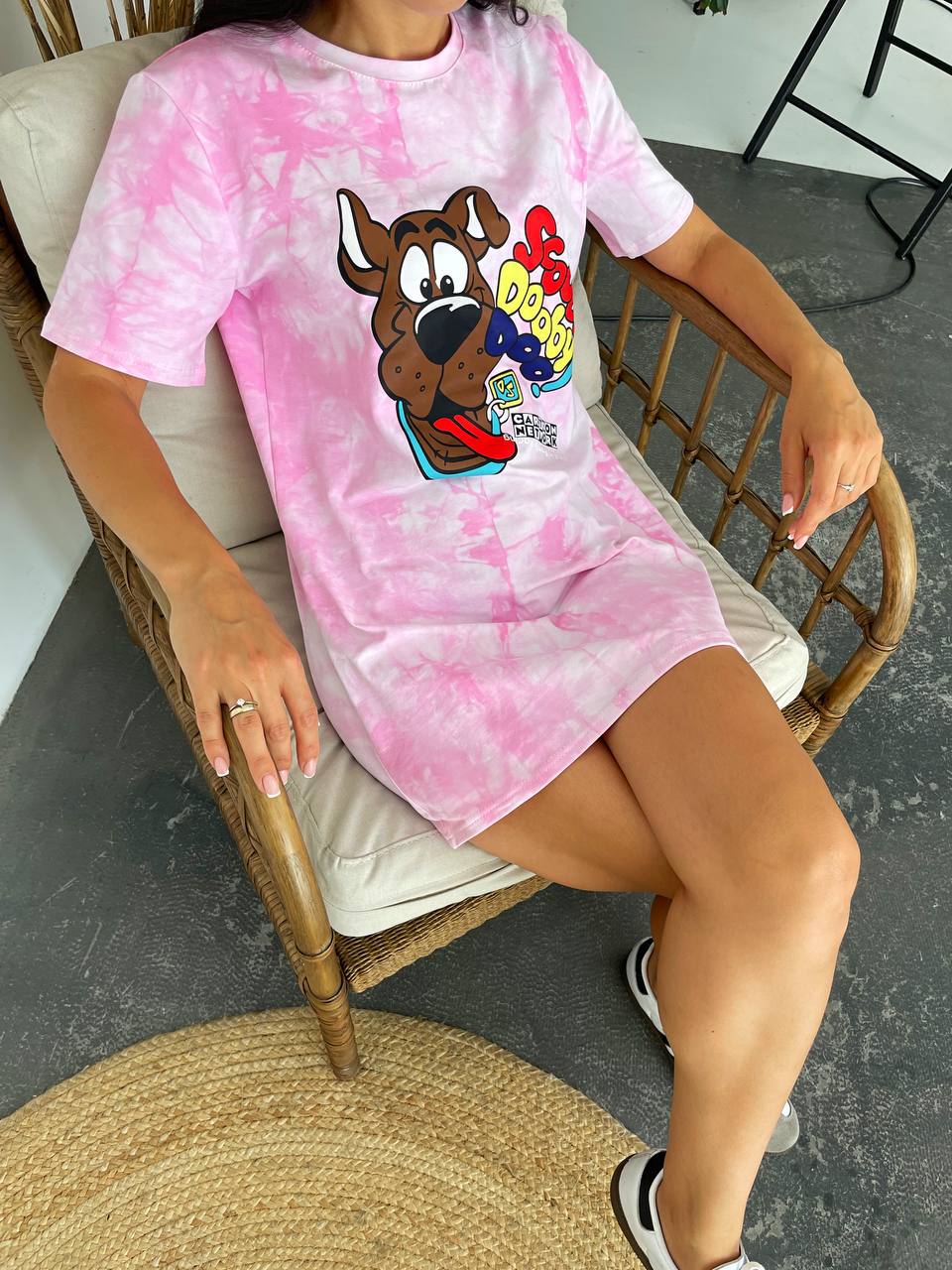 A woman sitting in a wicker chair wearing a pink and white tie-dye Scooby-Doo graphic t-shirt dress. The dress features a large cartoon graphic of Scooby-Doo holding a red cup with the text 'POO-DOO' on the front.