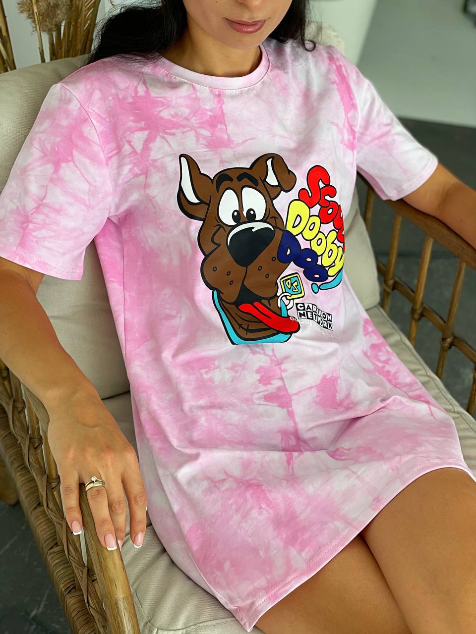 A woman wearing a pink and white tie-dye Scooby-Doo graphic t-shirt dress while sitting in a wicker chair.