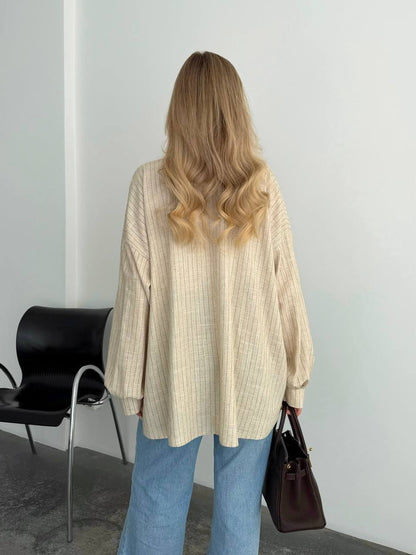 A woman with blonde wavy hair is shown from behind wearing a cream-colored ribbed knit sweater and light blue jeans, holding a dark brown handbag in a minimalist indoor setting.