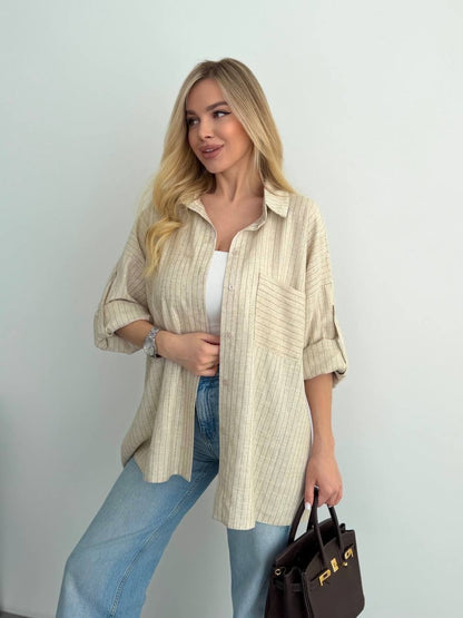 A woman models a light beige and white vertically striped button-down shirt with rolled-up sleeves, paired with light blue jeans and a white tank top, holding a dark brown handbag.
