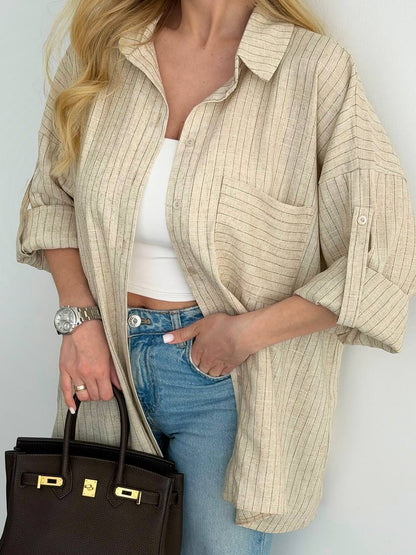 A woman wearing an oversized beige button-up shirt with thin vertical stripes, styled over a white tank top and blue jeans, holding a black handbag with gold hardware.