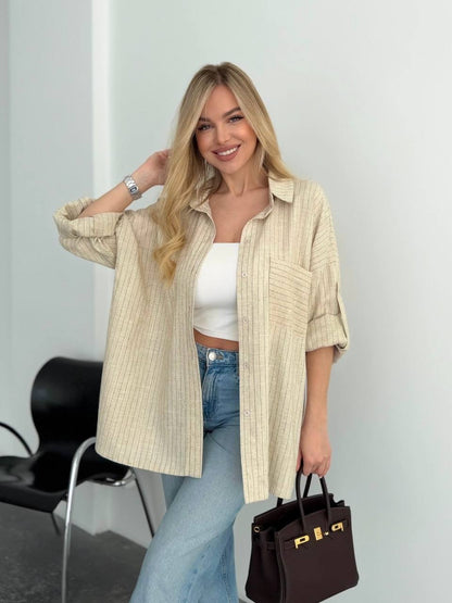 A woman models a light beige and white vertically striped button-up shirt over a white crop top and blue jeans, holding a dark brown handbag with gold lettering.
