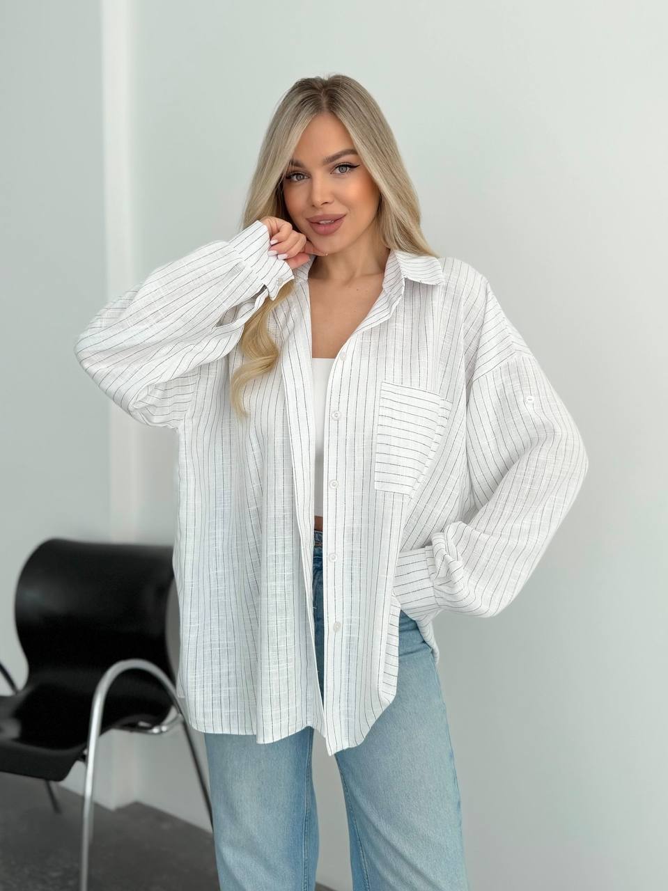 A woman modeling a white long-sleeve button-up shirt with thin vertical gray stripes, worn open over a white tank top and light blue jeans, standing in a minimalist indoor setting.