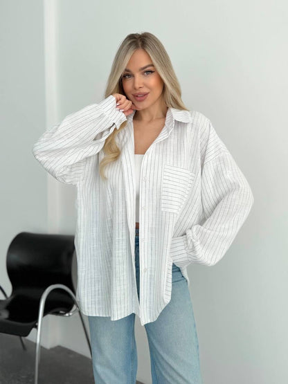 A woman modeling a white long-sleeve button-up shirt with thin vertical gray stripes, worn open over a white tank top and light blue jeans, standing in a minimalist indoor setting.
