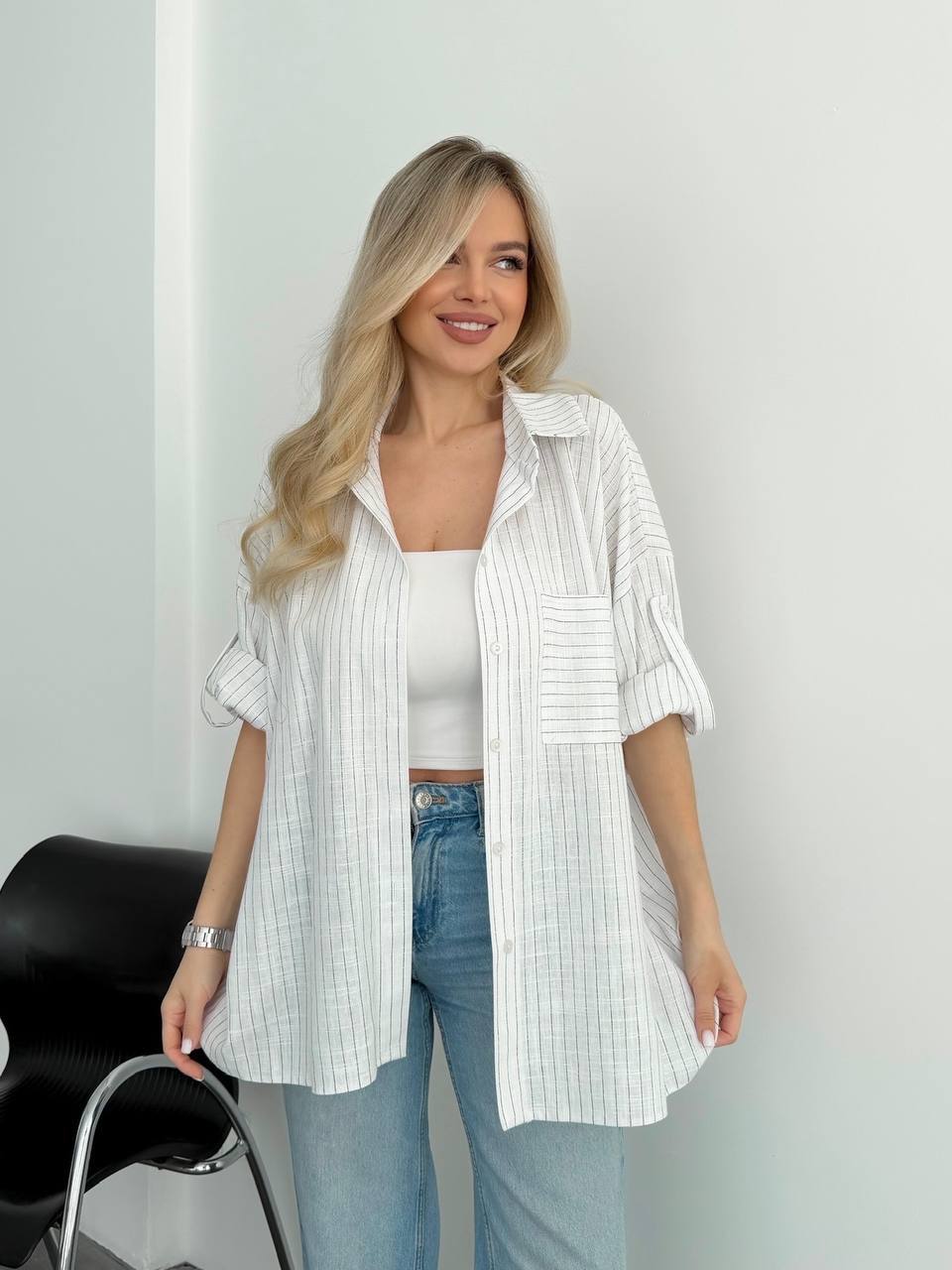 A woman wearing a white and light blue vertically striped button-down shirt over a white tank top and light-wash blue jeans, standing indoors against a white wall.