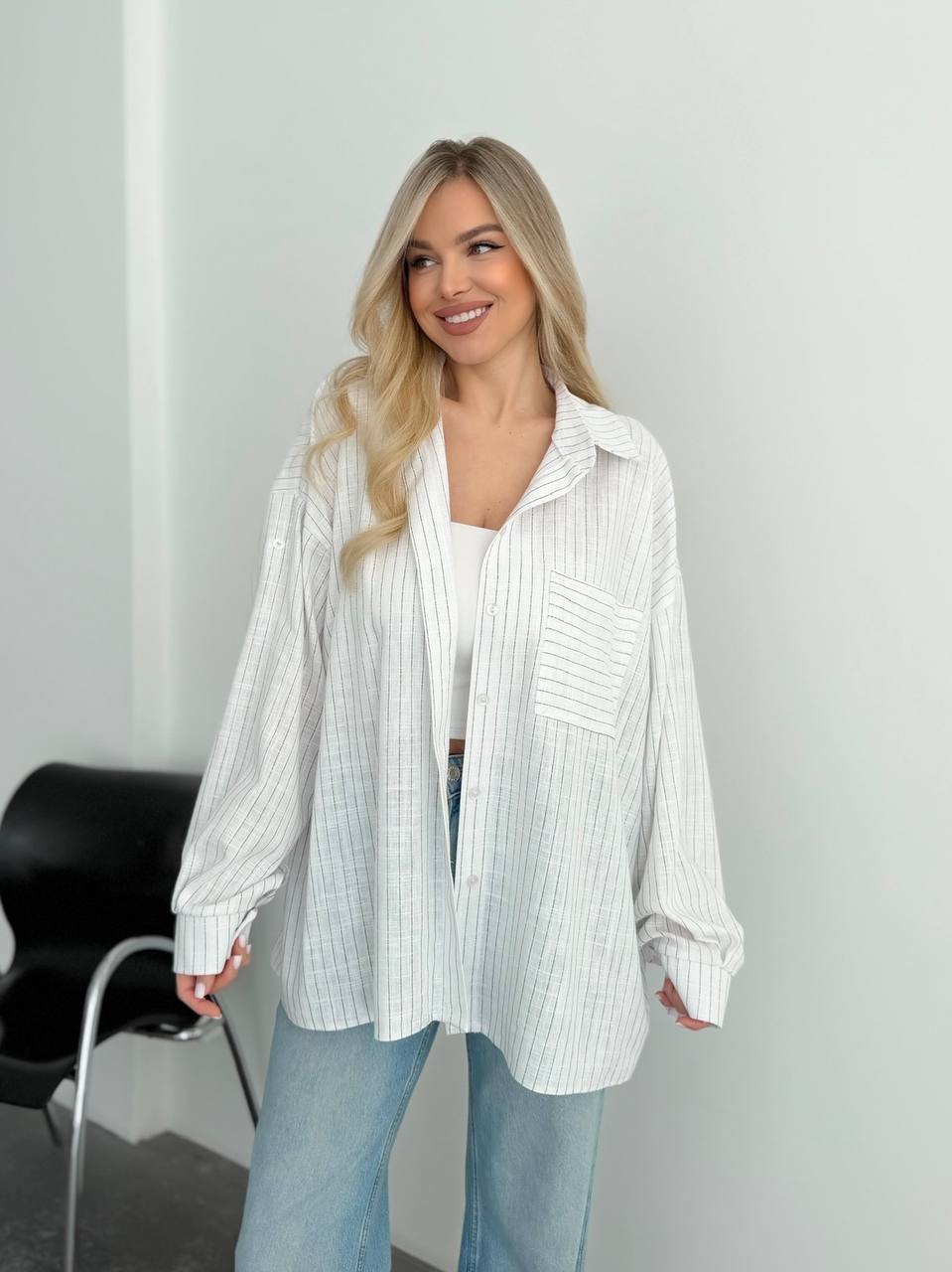 A woman wearing a white and light blue vertically striped button-up shirt over a white top and light blue jeans, standing in a minimalist room with a white wall and a black chair visible.