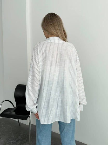 A woman viewed from behind wearing an oversized white button-up shirt with a subtle vertical stripe pattern and light blue jeans, standing in a minimalist room with a black chair against a white wall.