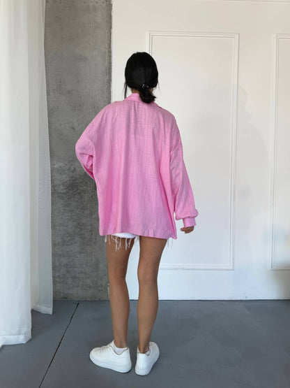 A person standing with their back to the camera wearing an oversized pink linen shirt, white shorts, and white sneakers in a minimalist room with a gray concrete wall and white door.