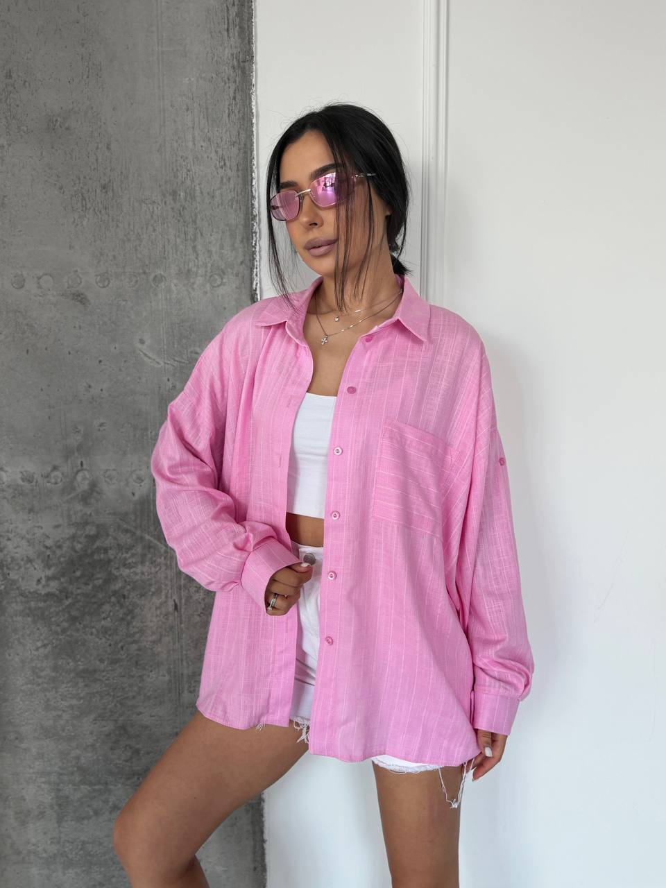 A woman wearing an oversized pink button-up shirt over a white crop top and white shorts, standing against a split gray and white wall.