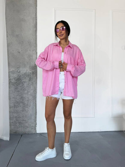 A woman modeling an oversized pink striped button-up shirt, styled with white shorts and white sneakers, standing in front of a white paneled wall.