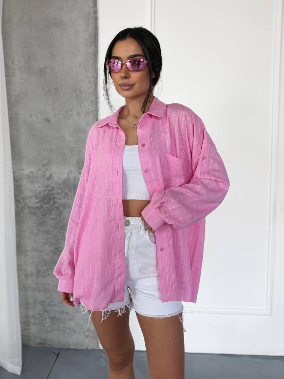 A woman modeling a pink textured button-up shirt over a white crop top and white frayed denim shorts, standing against a white paneled wall with a gray concrete accent.