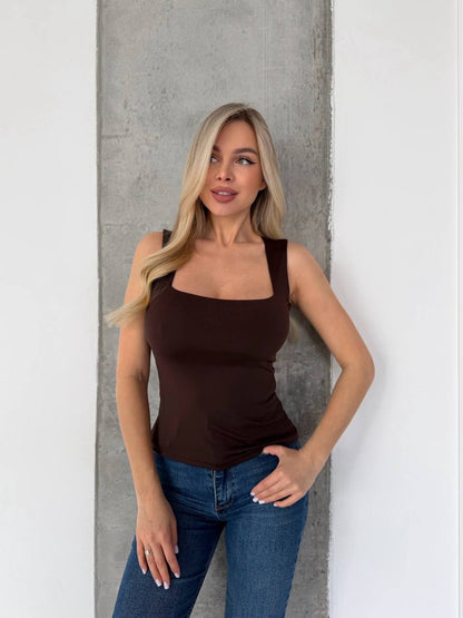 A blonde woman wearing a fitted dark brown scoop-neck tank top and blue jeans, posing against a vertical concrete pillar between white walls.