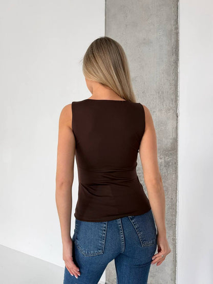 A woman viewed from behind wearing a fitted sleeveless dark brown top and blue jeans, standing against a white wall with a concrete pillar.