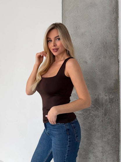 A blonde woman wearing a dark brown scoop-neck tank top and blue jeans, posing against a split white and gray concrete wall.
