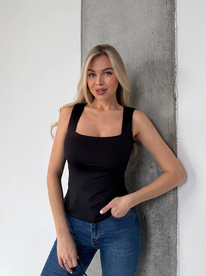 A blonde woman wearing a black square-neck tank top and blue jeans, posing against a white wall with a textured gray concrete pillar.
