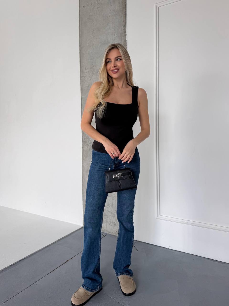 A woman stands in a minimalist white room with a concrete pillar, wearing a black tank top, blue jeans, and beige slip-on shoes, while holding a small black handbag with a silver logo.