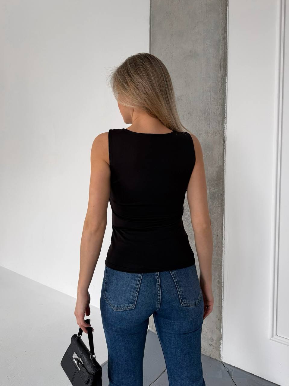 A woman viewed from behind wearing a fitted black sleeveless top and blue jeans, standing indoors against a white wall and concrete pillar, holding a black handbag.