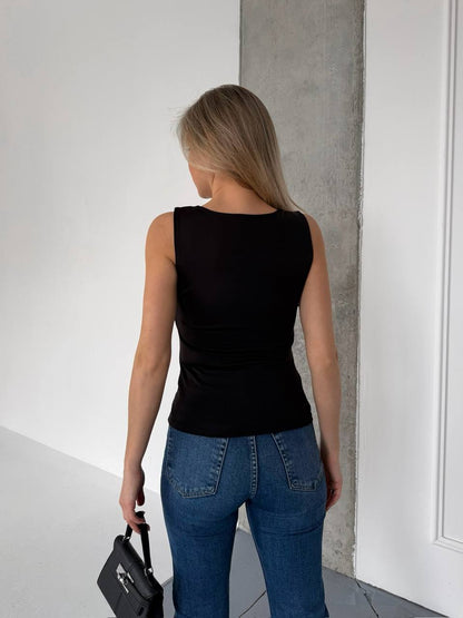 A woman viewed from behind wearing a fitted black sleeveless top and blue jeans, standing indoors against a white wall and concrete pillar, holding a black handbag.