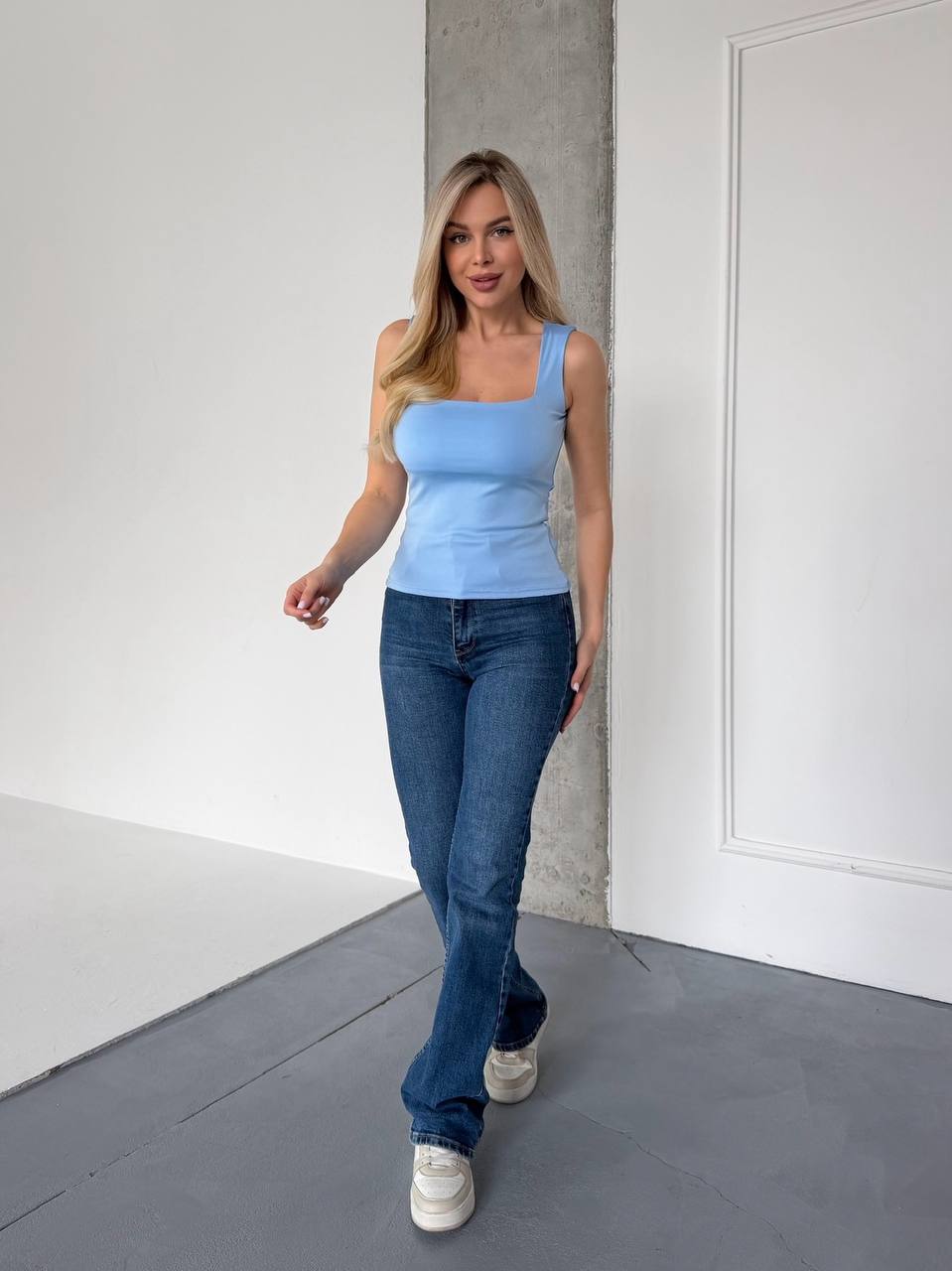 A woman modeling a light blue ribbed tank top and dark blue jeans in a minimalist studio setting with white walls and a gray floor.