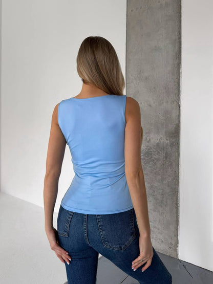 A woman viewed from behind wearing a light blue sleeveless top and dark blue jeans, standing in a minimalist room with white walls and a concrete pillar.