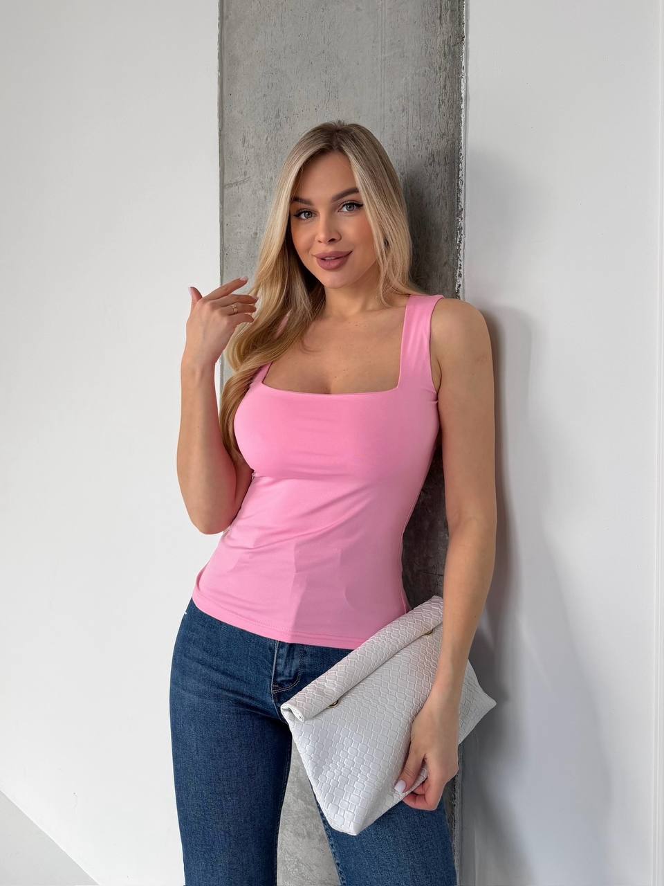 A woman with blonde hair wearing a bright pink tank top and blue jeans, leaning against a gray concrete pillar next to a white wall while holding a textured white clutch.