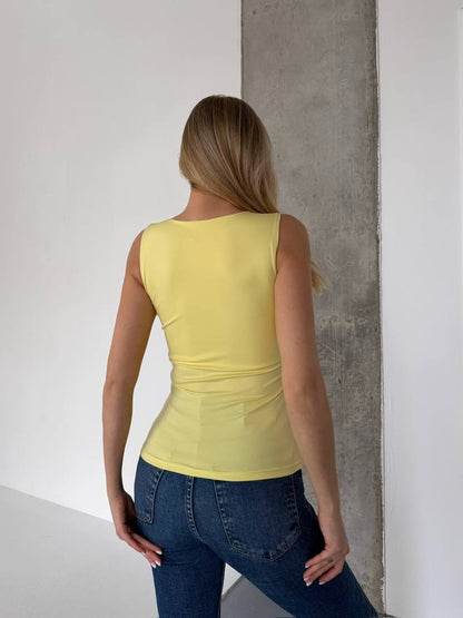 A woman viewed from behind wearing a fitted sleeveless yellow top and blue jeans, standing in a minimalist room with white walls and a concrete pillar.