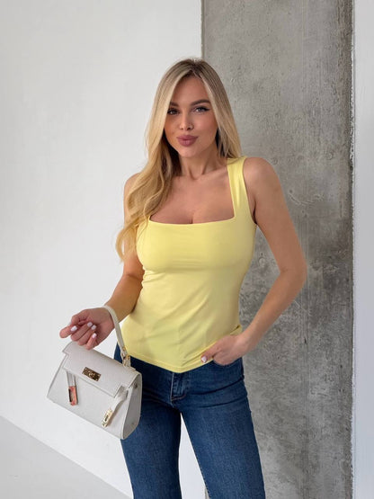 A woman modeling a pale yellow sleeveless square-neck tank top with blue jeans, holding a small white handbag against a split white and gray wall background.