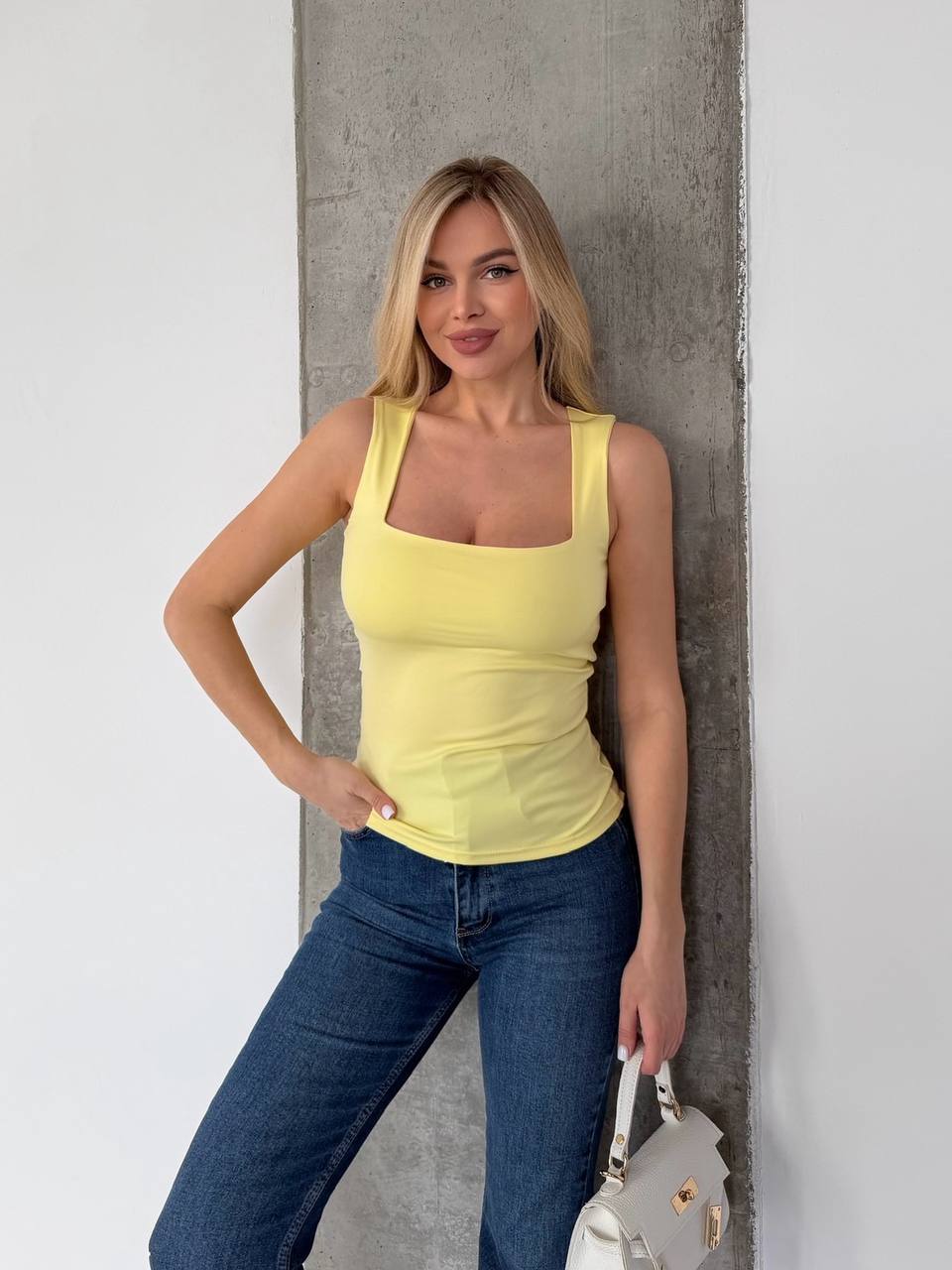 A woman wearing a fitted yellow tank top and blue jeans stands against a gray concrete pillar, holding a small white handbag.