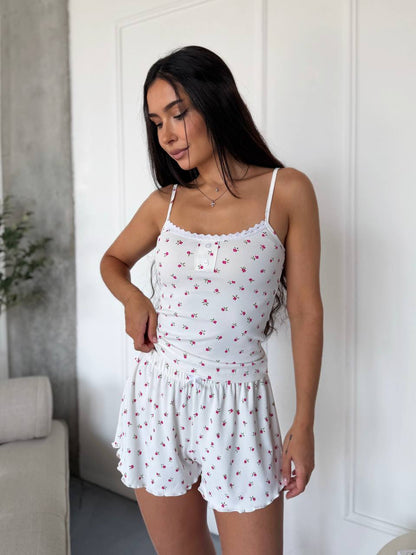 A woman modeling a white camisole and shorts set with a small red cherry print, standing in a brightly lit room with white paneled walls.