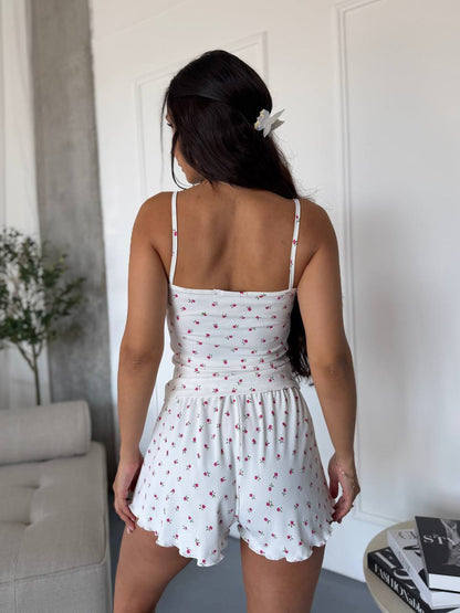 A woman viewed from behind wearing a white sleeveless romper with thin straps and a small red floral print, standing indoors against a white paneled wall.