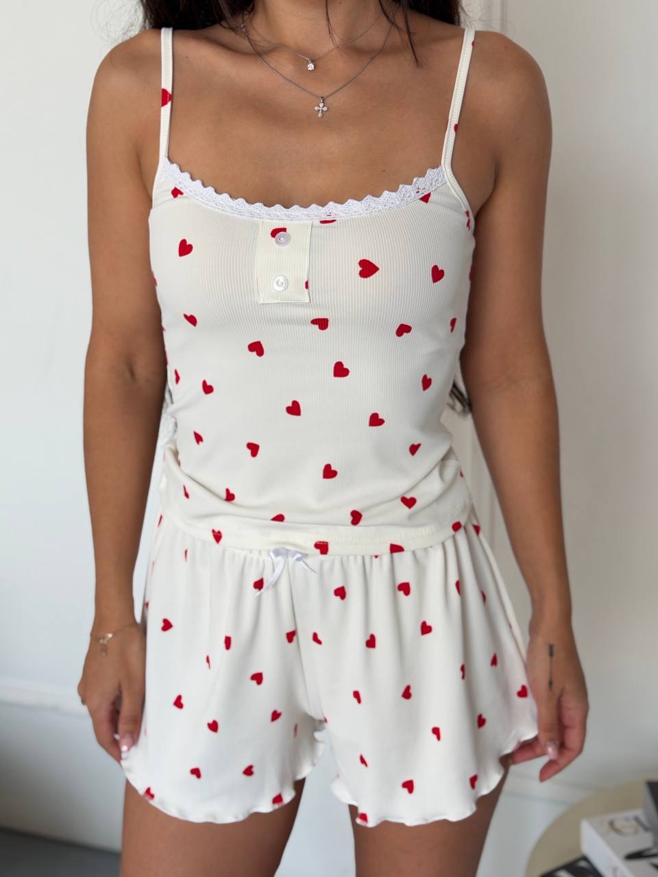 A woman wearing a white ribbed camisole top and matching shorts set with a red heart print, lace trim, and a white drawstring waist.
