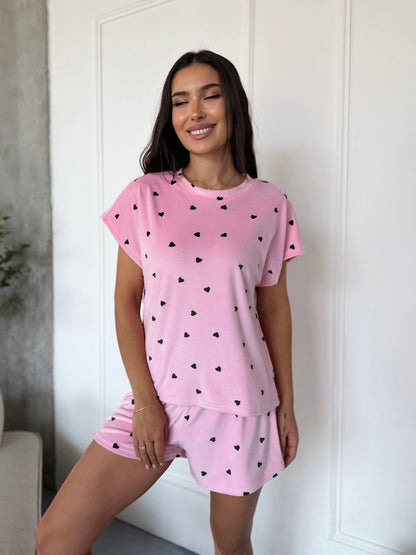 A woman modeling a pink two-piece pajama set with a small black heart pattern, consisting of a short-sleeve t-shirt and matching shorts, standing in front of a white paneled door.