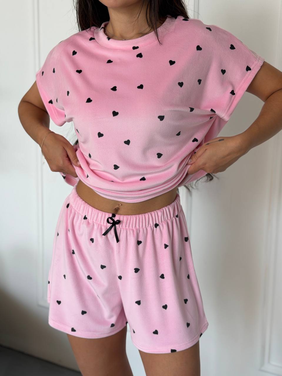 A woman modeling a pink two-piece pajama set with small black heart prints. The set includes a short-sleeve cropped t-shirt and matching drawstring shorts, shown against a white paneled door background.