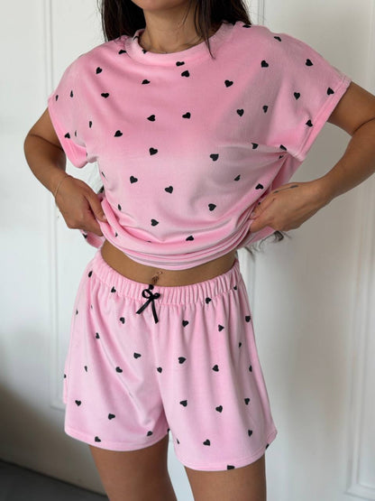 A woman modeling a pink two-piece pajama set with small black heart prints. The set includes a short-sleeve cropped t-shirt and matching drawstring shorts, shown against a white paneled door background.