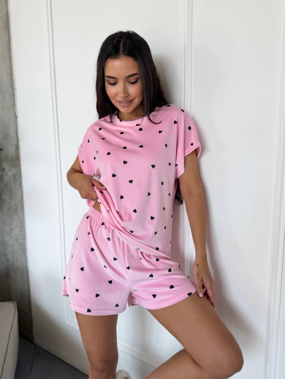 A woman modeling a pink two-piece lounge set consisting of a short-sleeve t-shirt and matching shorts, both printed with small black hearts, posed against a white paneled wall.