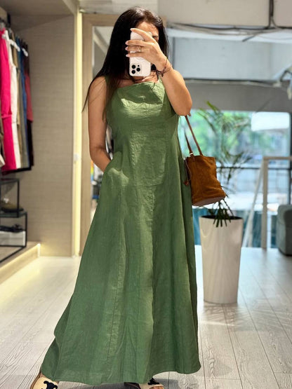 A woman takes a mirror selfie in a boutique wearing a sleeveless, floor-length sage green dress. She is holding a brown leather shoulder bag and wearing a white phone to take the photo. The boutique has light-colored walls and wooden floors.